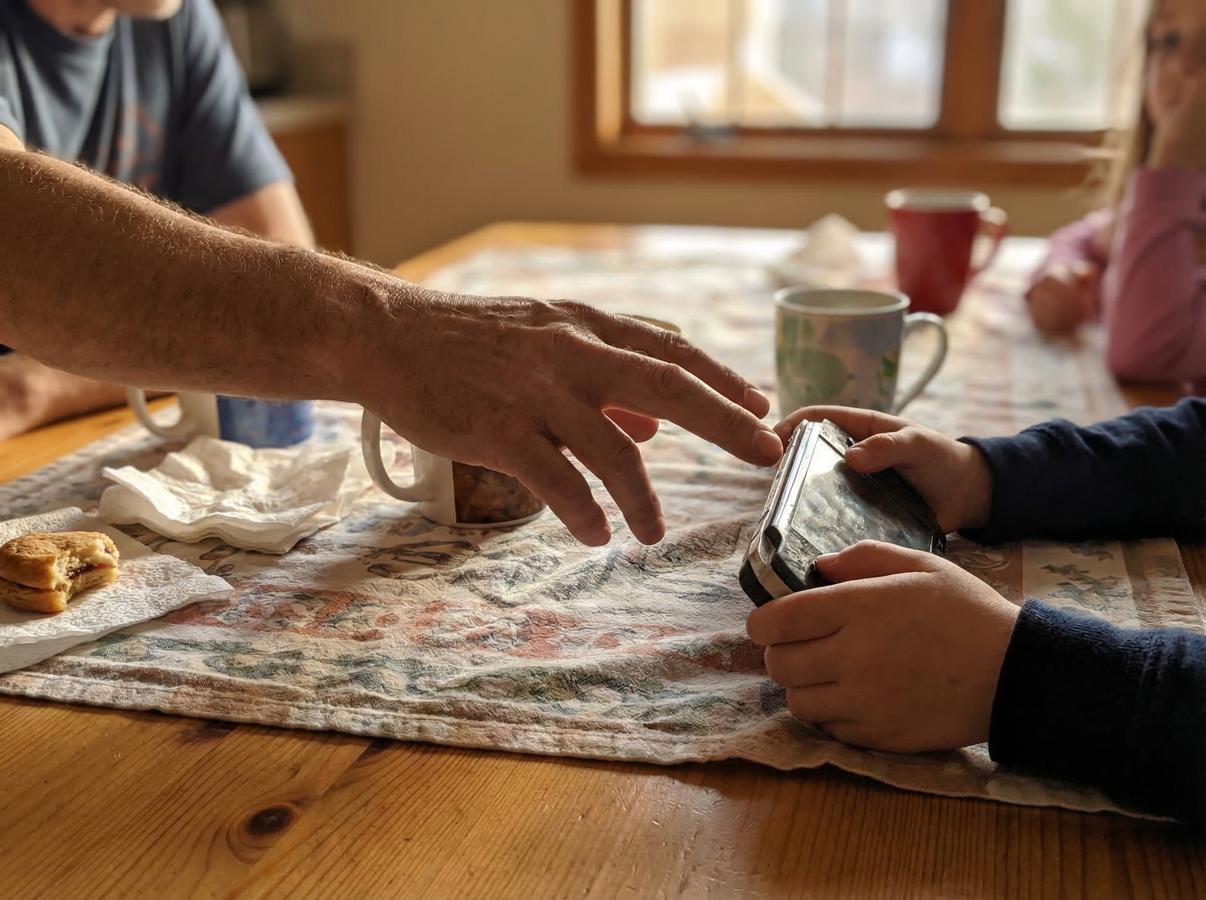A kitchen table scene with a parent's hand reaching toward a gaming device while a child clutches it protectively, showing te
