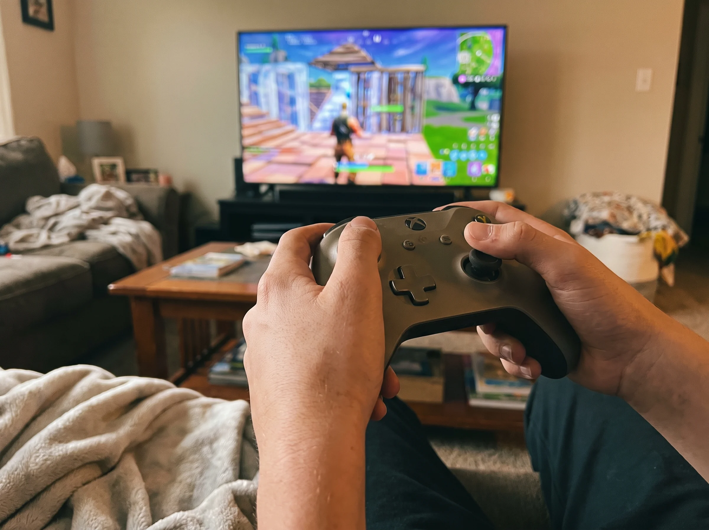 Close-up of a teen boy's hands on a game controller with a Fortnite-style match on the TV screen in the background