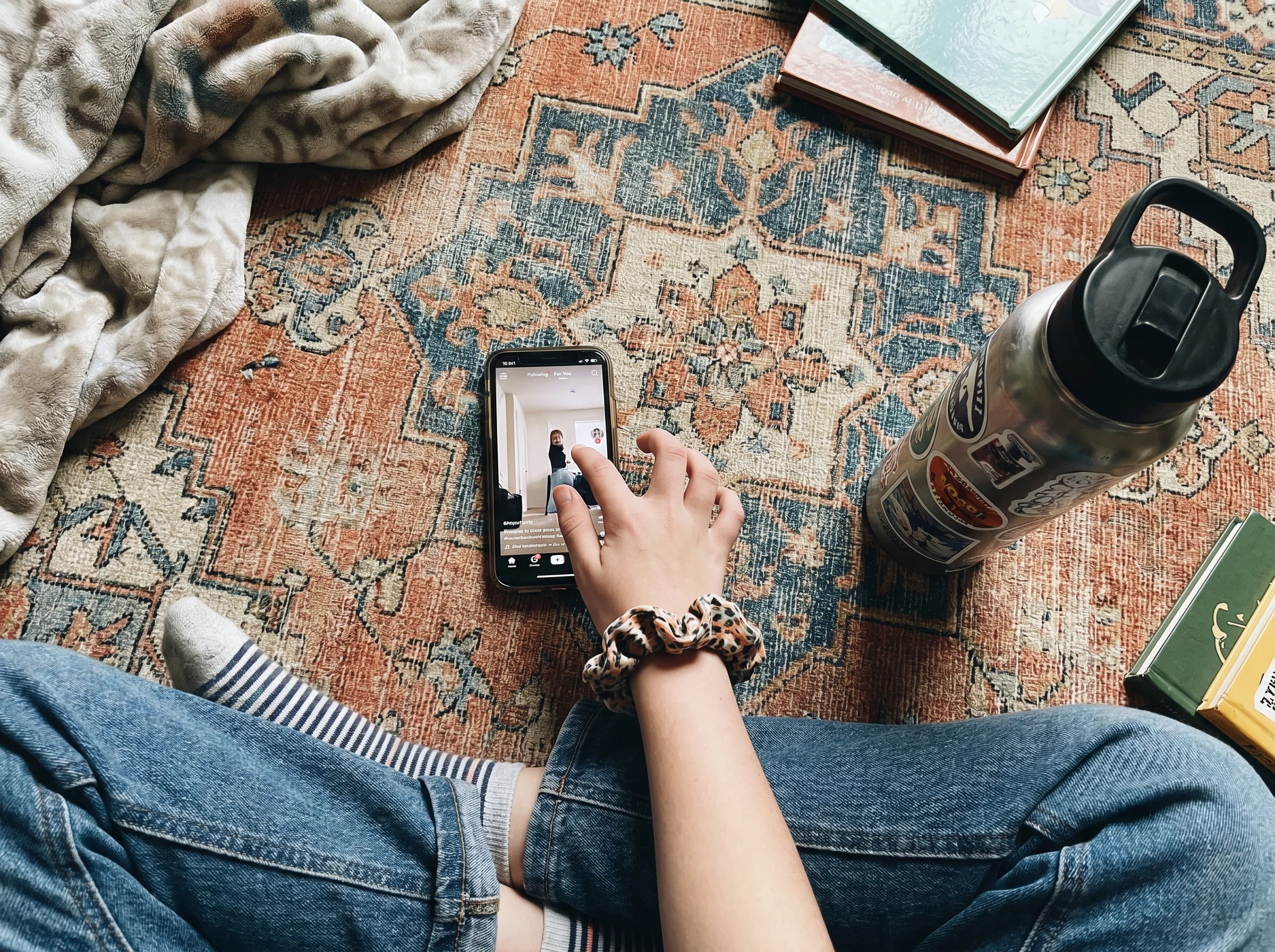 Overhead shot of a tween's hand scrolling a vertical video feed on a phone, sitting cross-legged on a bedroom rug with a wate