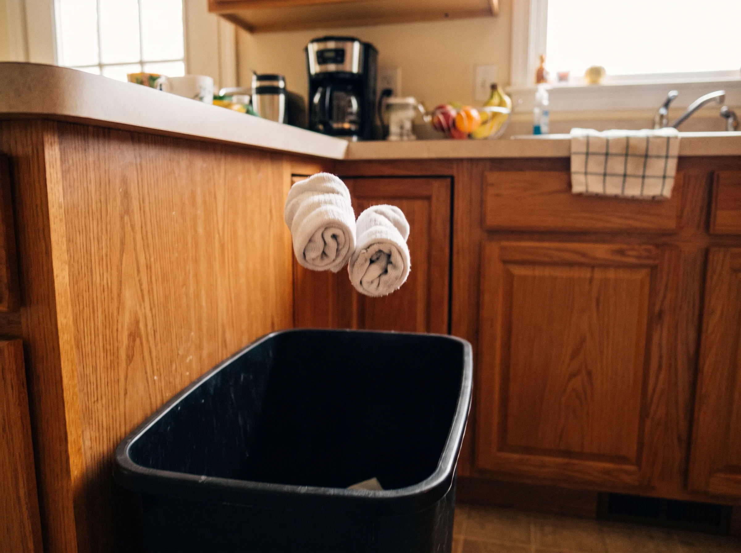 A rolled-up pair of white socks mid-air about to land in a black kitchen trash can, kitchen counter in the background