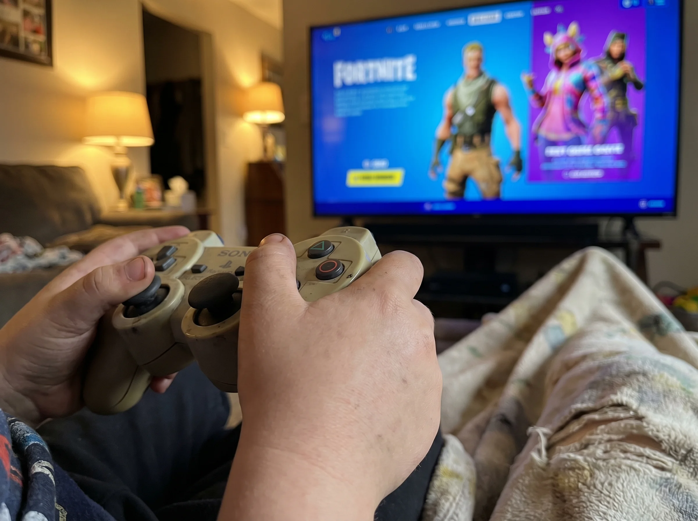 Close-up of a kid's hands holding a game controller, with a TV screen in the background showing a Fortnite character in defau