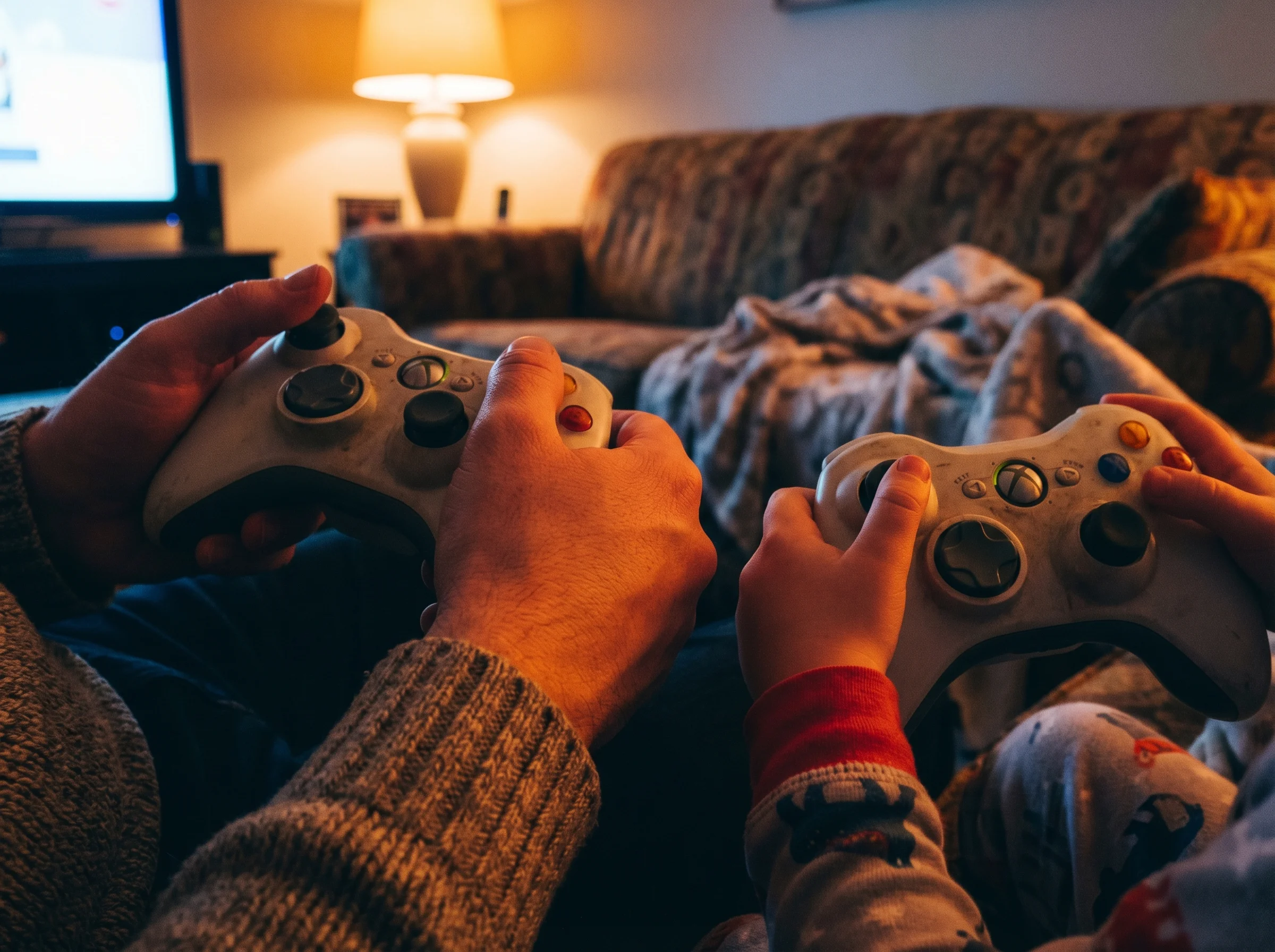 Close-up of parent and child's hands both holding game controllers, sitting close together with warm lighting suggesting even