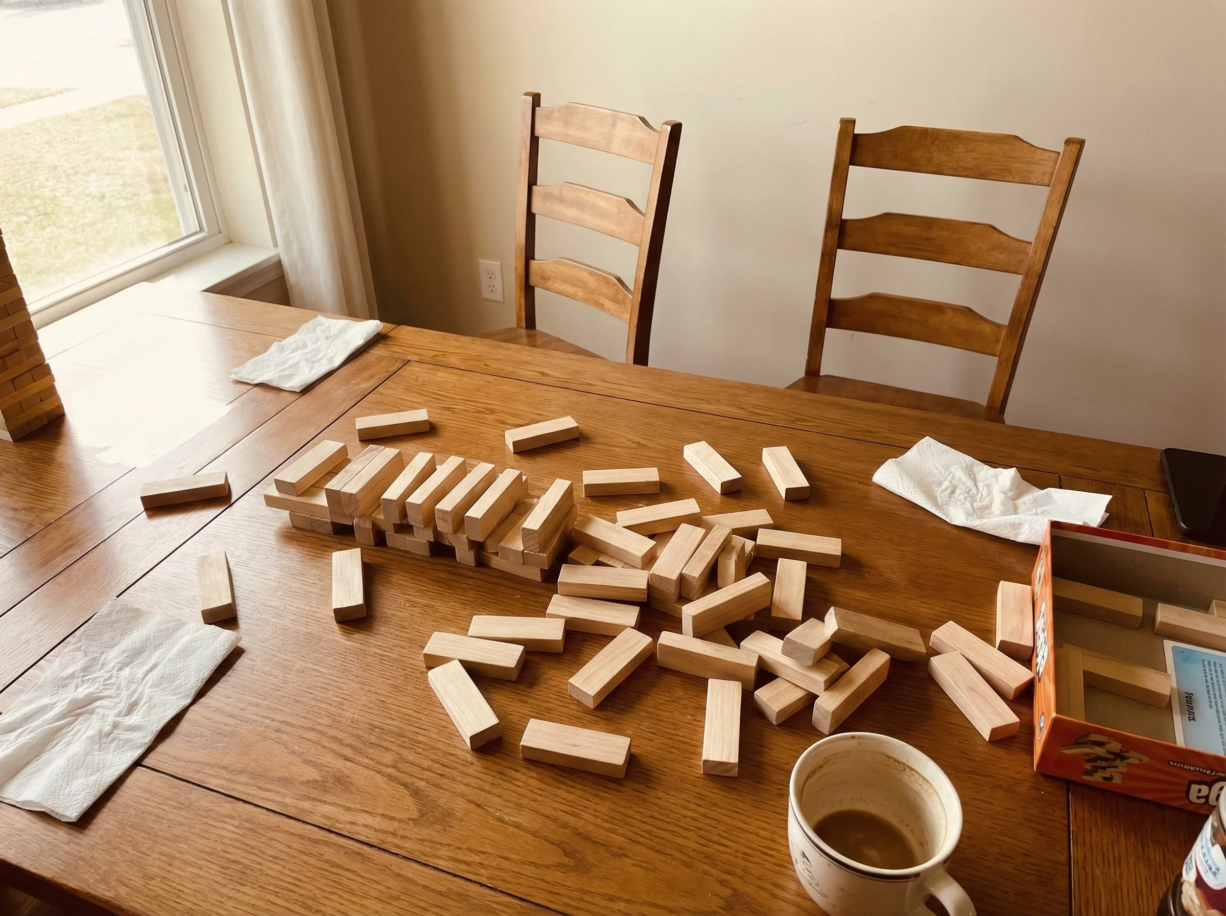 A toppled Jenga tower on a dining table with game pieces scattered, two empty chairs pushed back suggesting recent departure