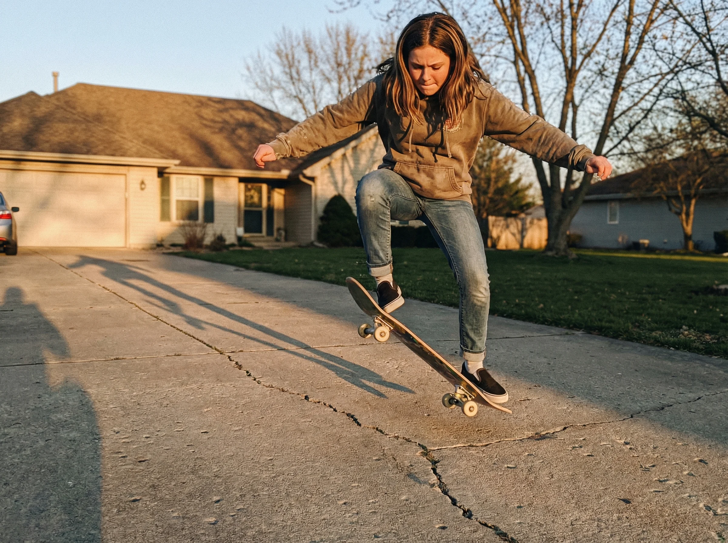 A teenage girl in the driveway with a skateboard, mid-attempt at an ollie, shot from ground level showing determination on he