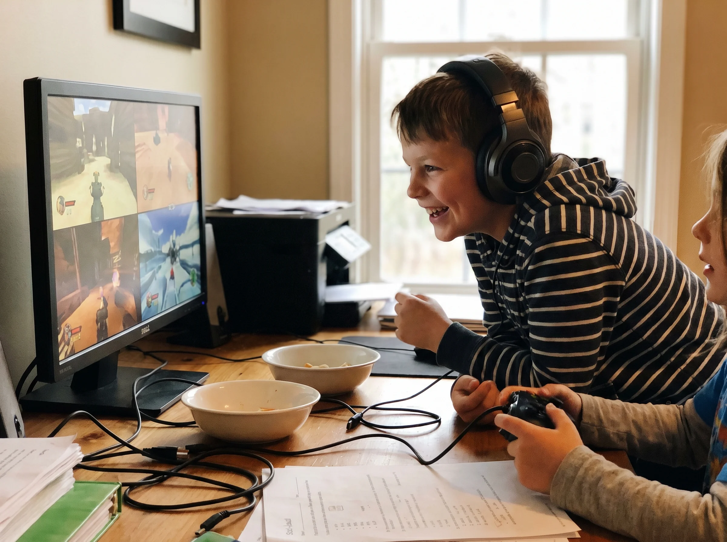 A boy wearing headphones at a desk, leaning forward and smiling at the screen, clearly engaged in a co-op game with a friend