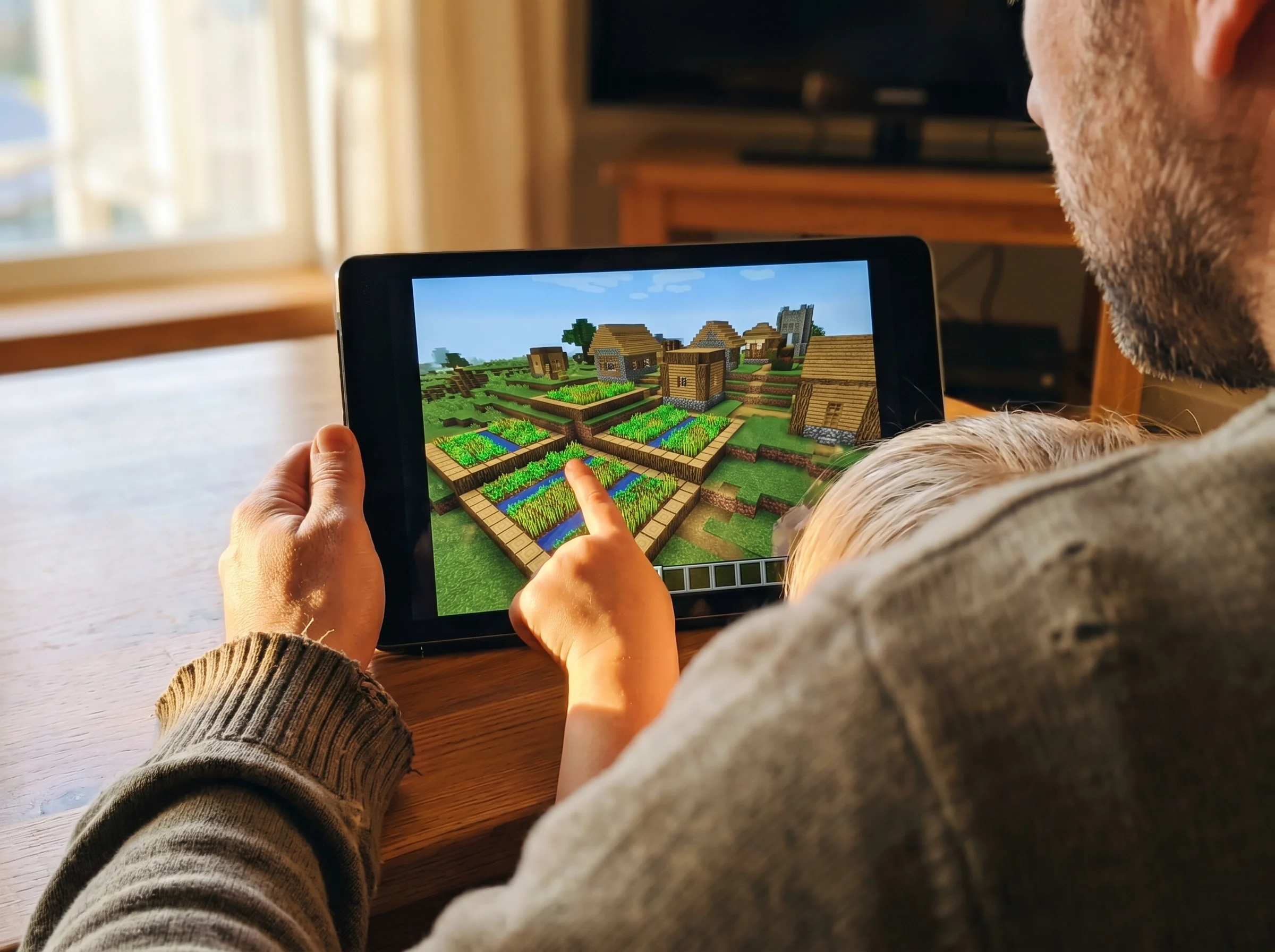 Close-up of a Minecraft world on a screen showing an elaborate village build with farms and structures, kid's hand pointing a