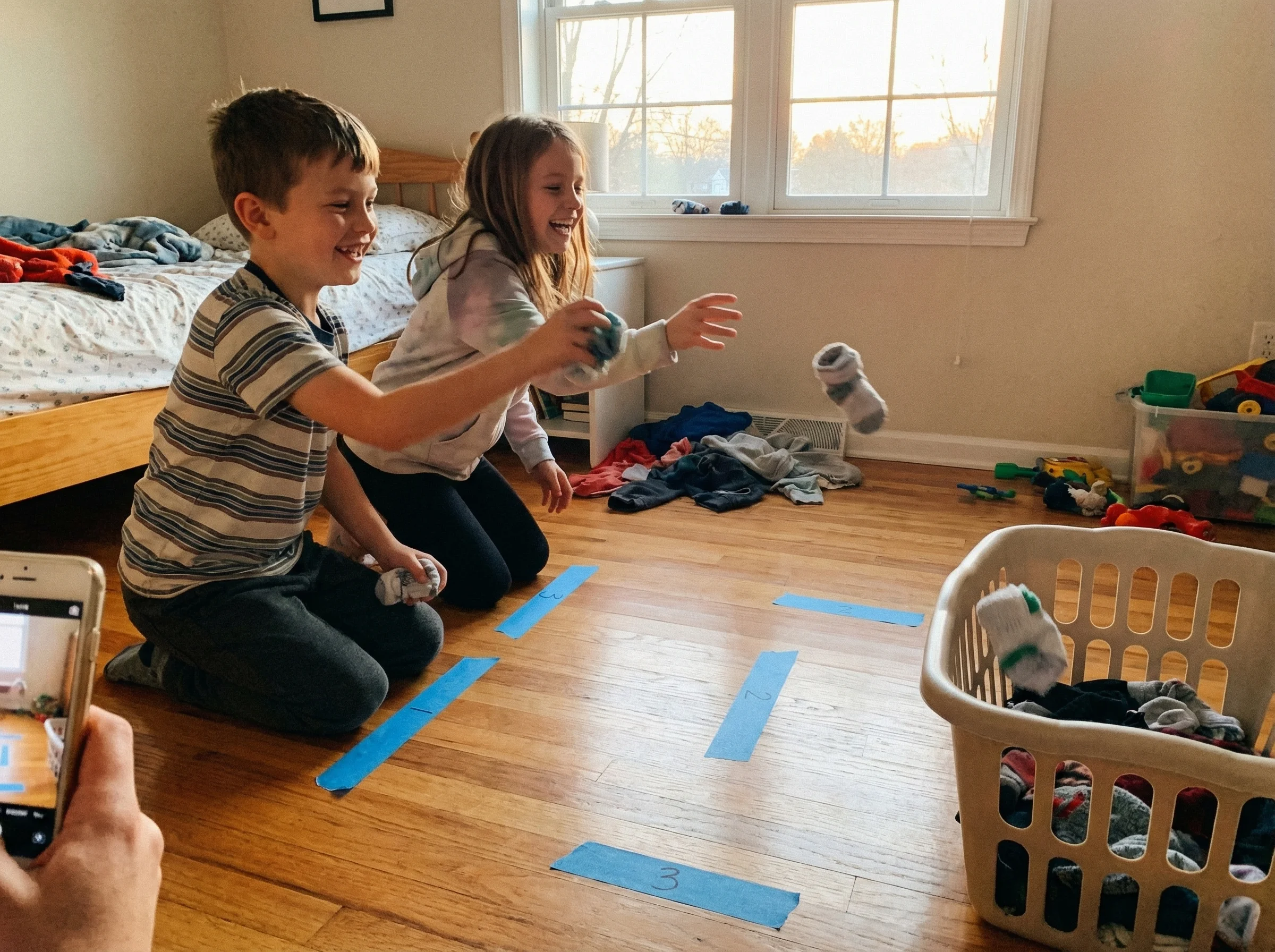 Kids tossing rolled-up socks into a laundry basket from different distances marked with masking tape on a bedroom floor