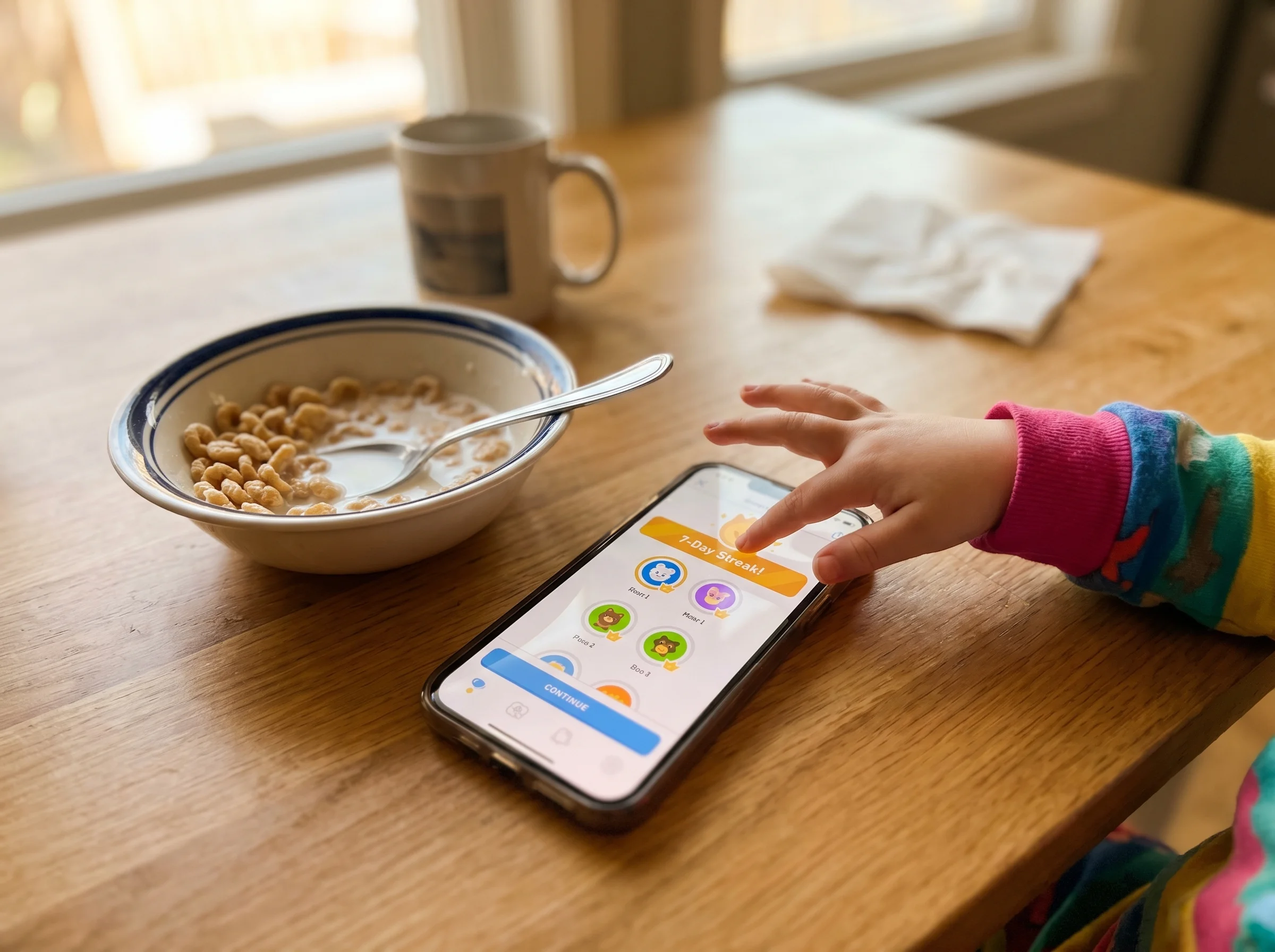 Phone showing a language learning app with a streak counter, kid's hand on the screen, breakfast cereal bowl nearby on a kitc