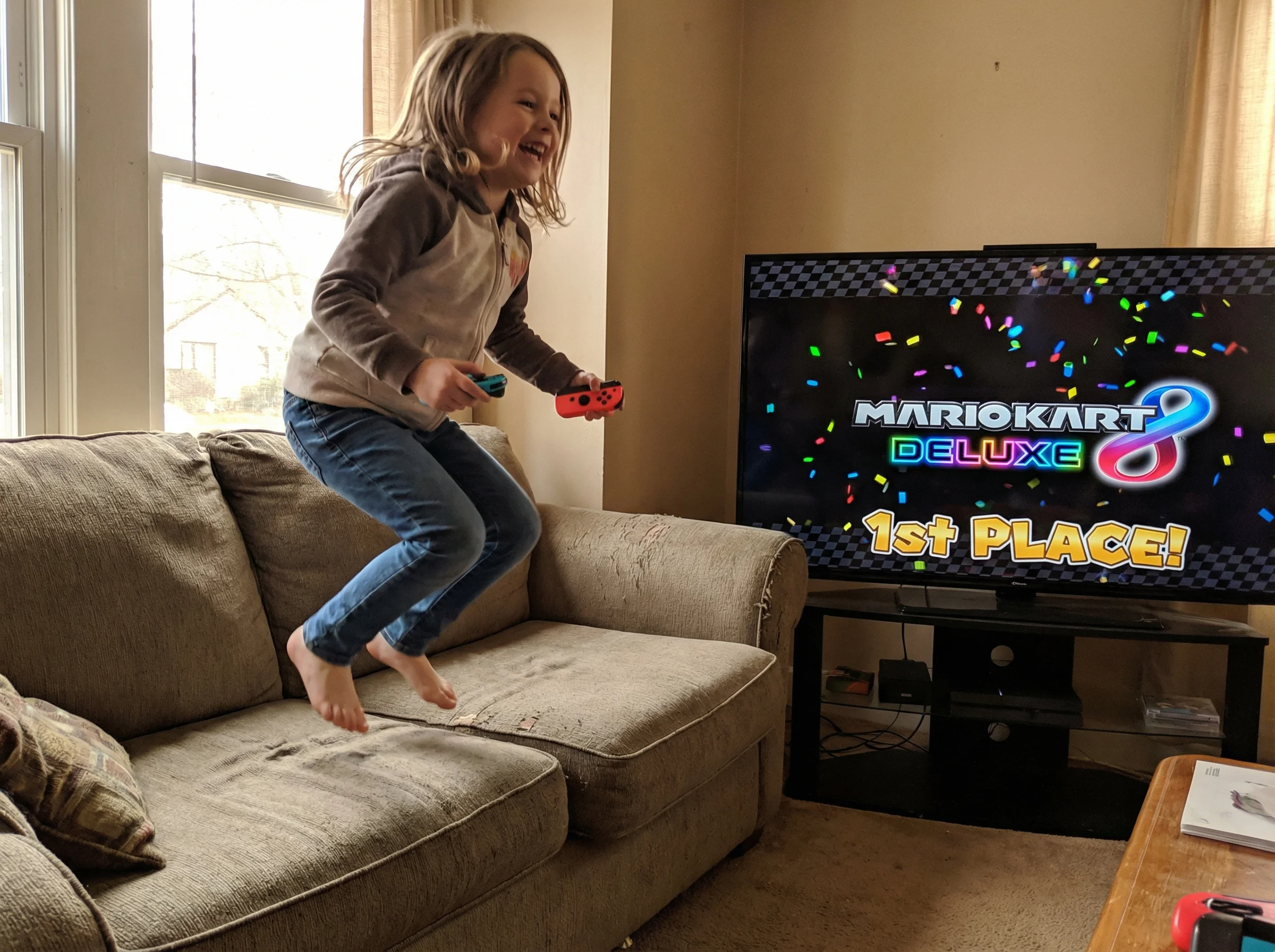 A girl jumping up from the couch in celebration, Nintendo Switch controller in hand, TV showing Mario Kart victory screen in