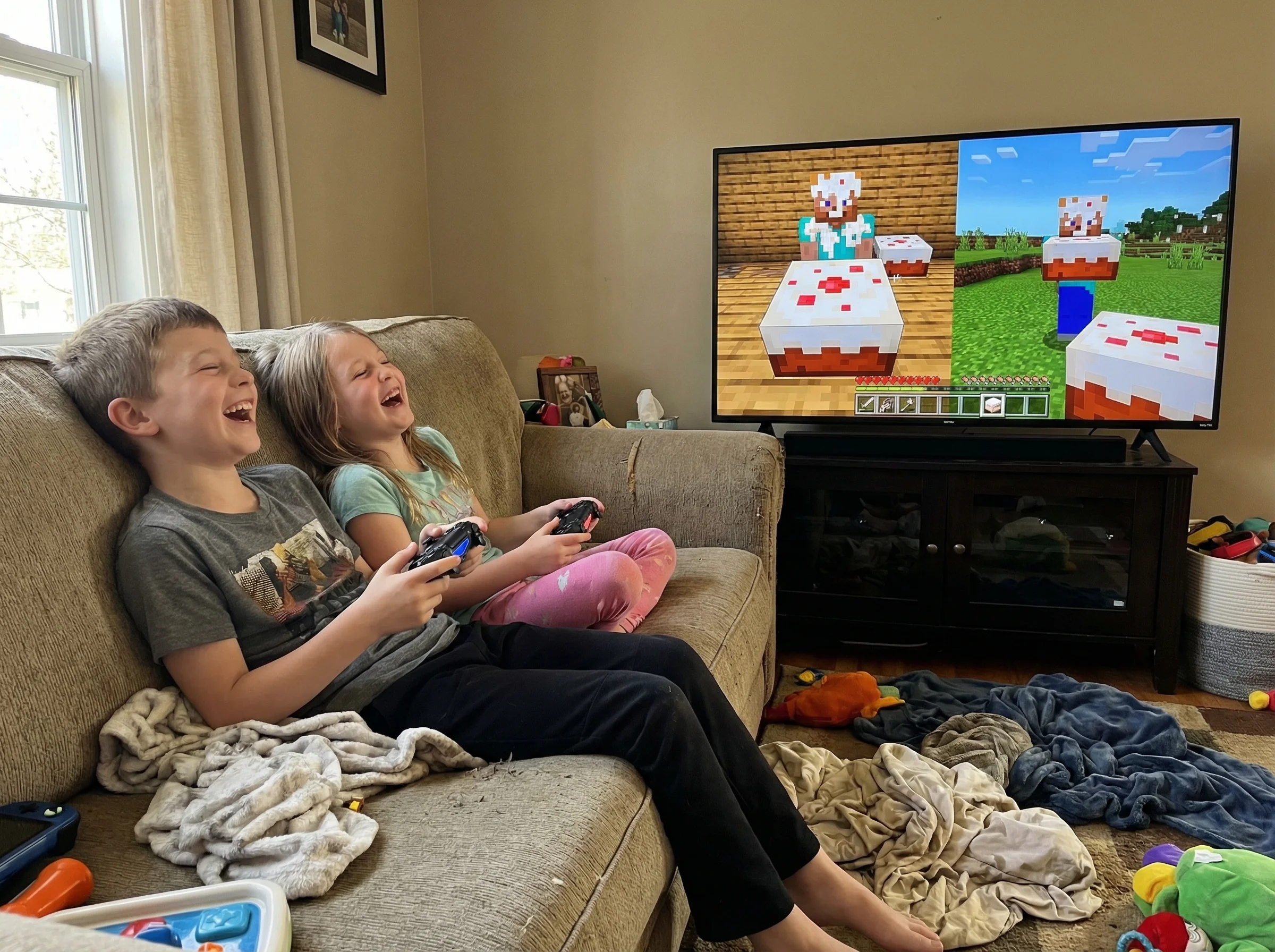 Two kids laughing together on a couch, game controllers in hand, split-screen showing playful Minecraft pranks with cake ever