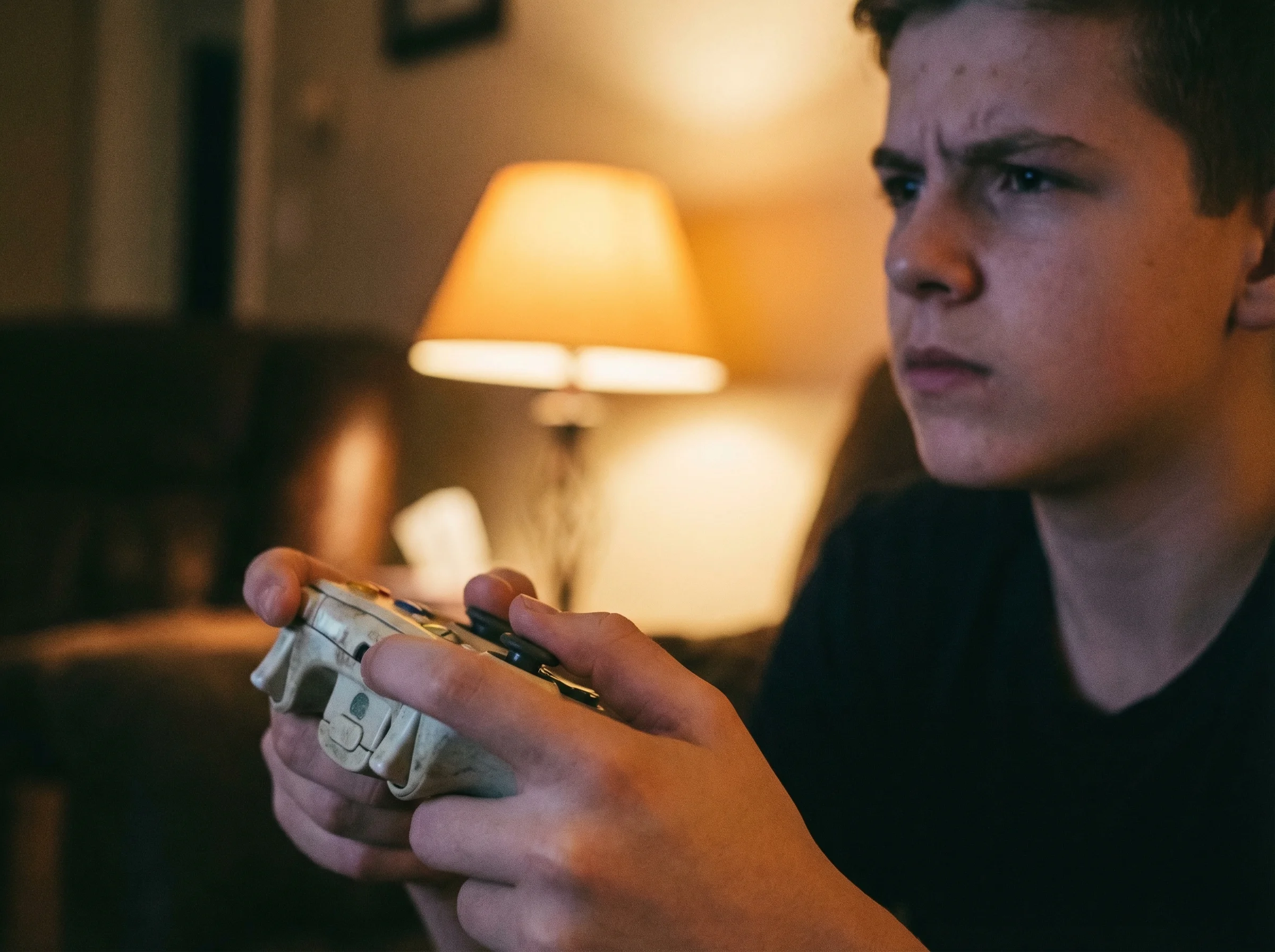 Close-up of a teenager's hands holding a game controller, focused expression visible in background, warm lamp lighting