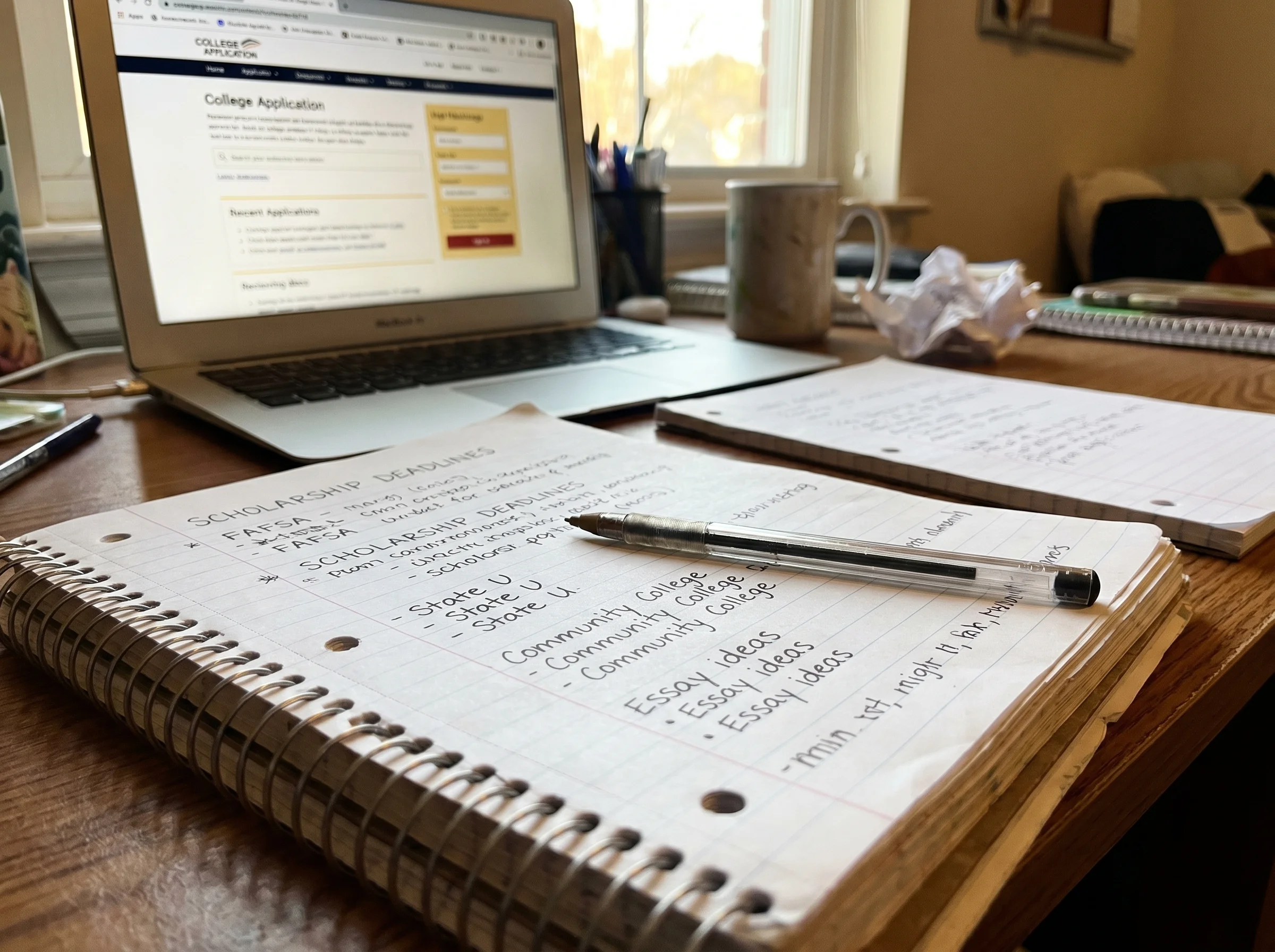Notebook on a desk with handwritten notes about scholarships and college names, pen resting on top, laptop slightly out of fo
