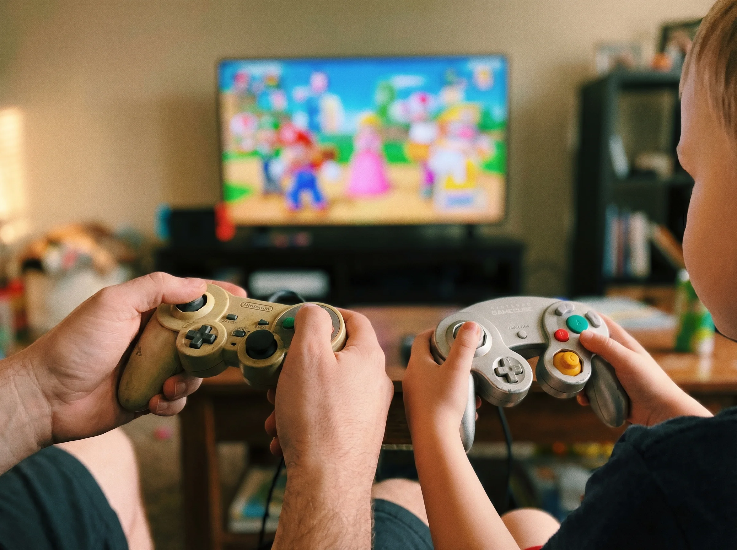 Close-up of two sets of hands holding game controllers, one adult and one child, with a blurred TV screen showing a colorful