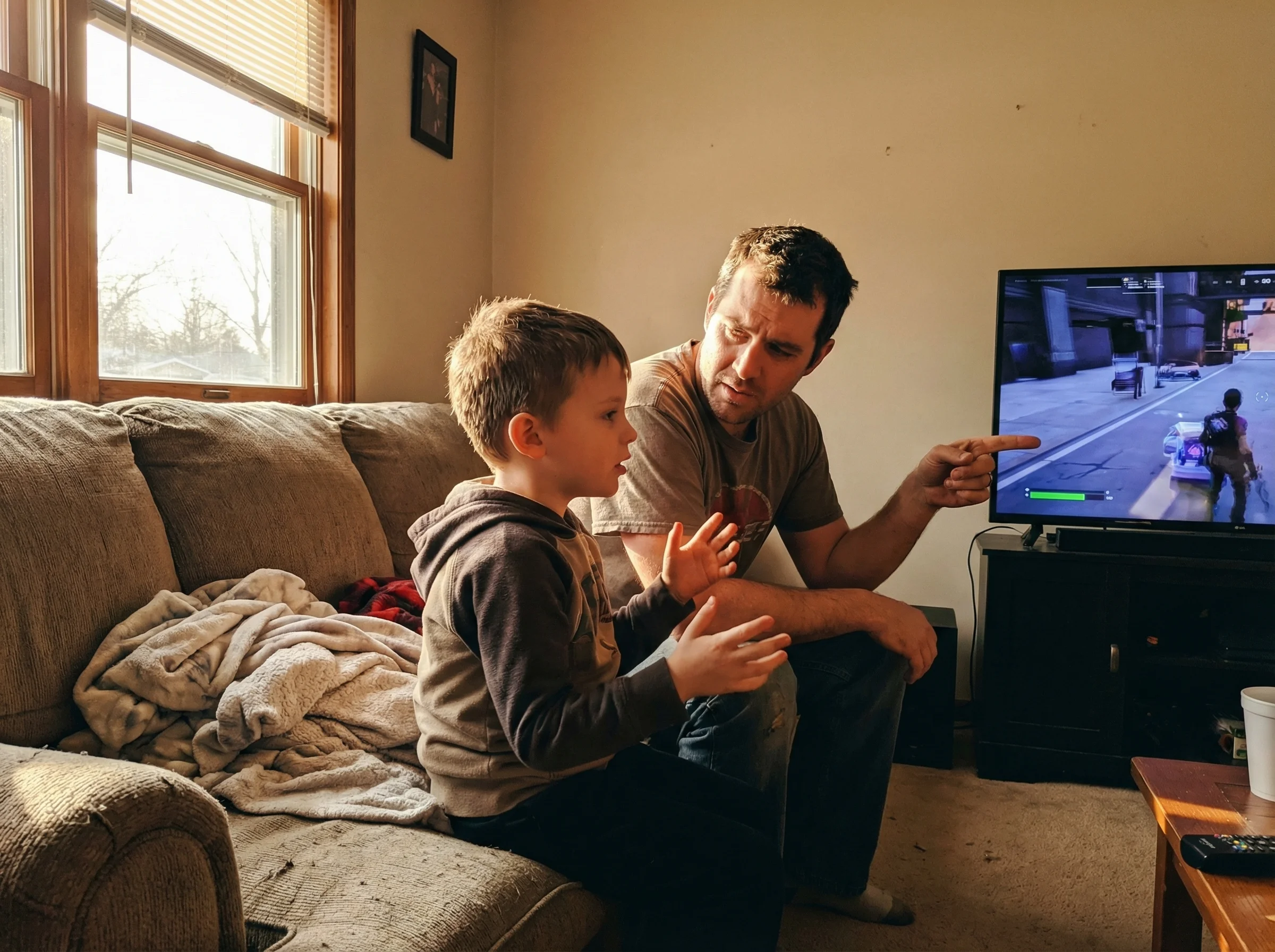 A dad sitting beside his son on the couch, both looking at the game screen, the dad asking a question while the son explains