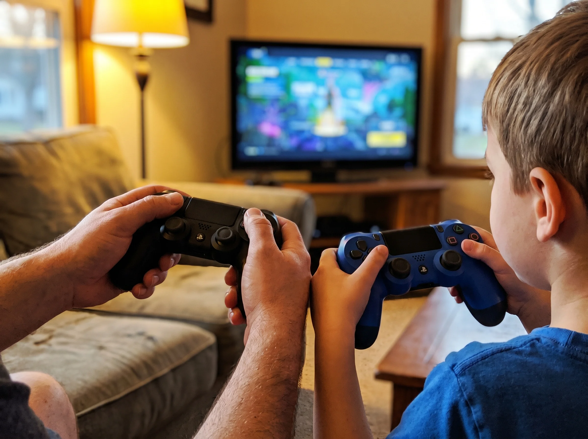 A close-up of a dad and son's hands both holding game controllers, side by side, the TV screen slightly out of focus in the b