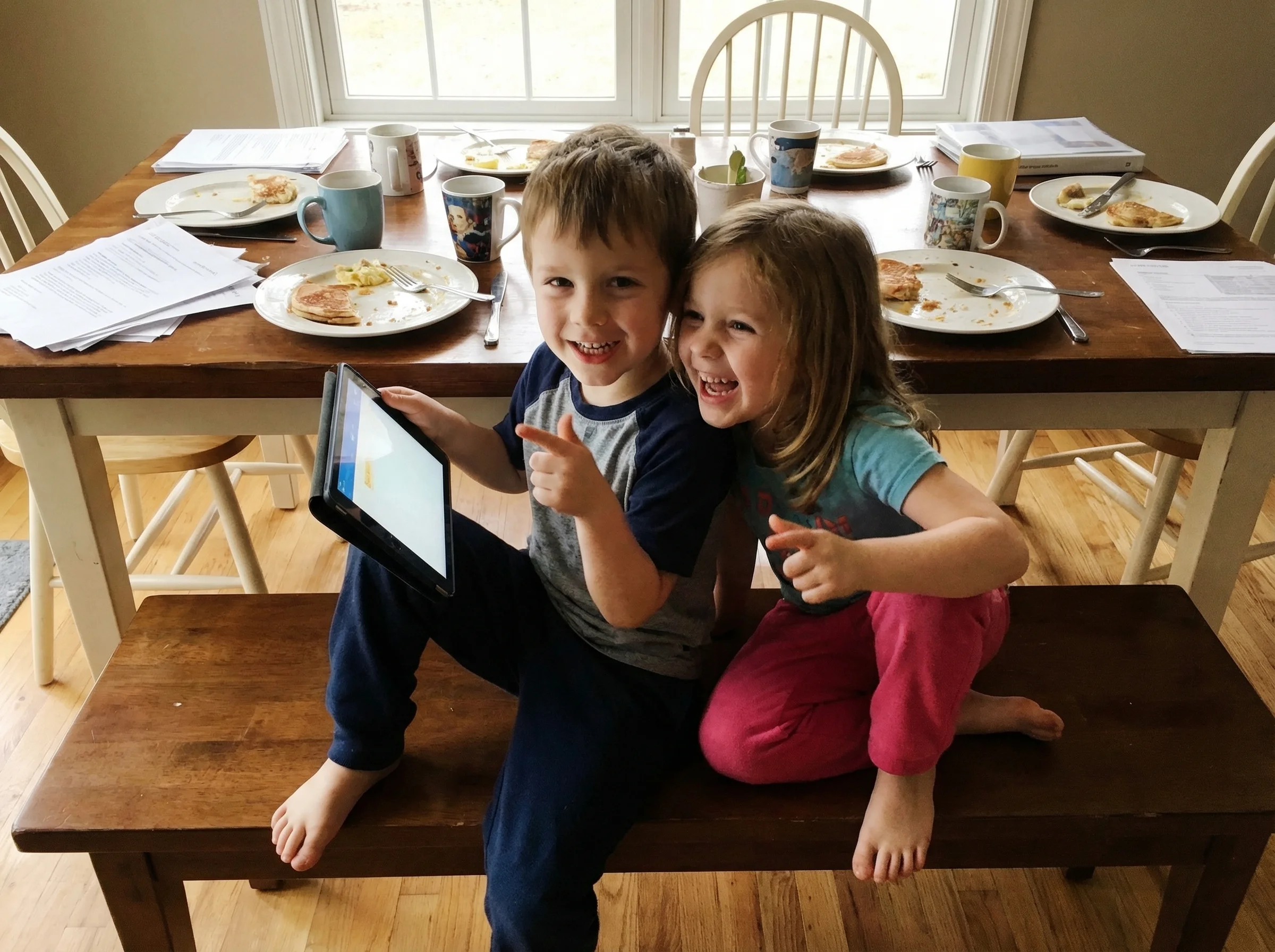 Two kids sitting side by side at a dining table, one showing the other something exciting on their tablet screen, both smilin