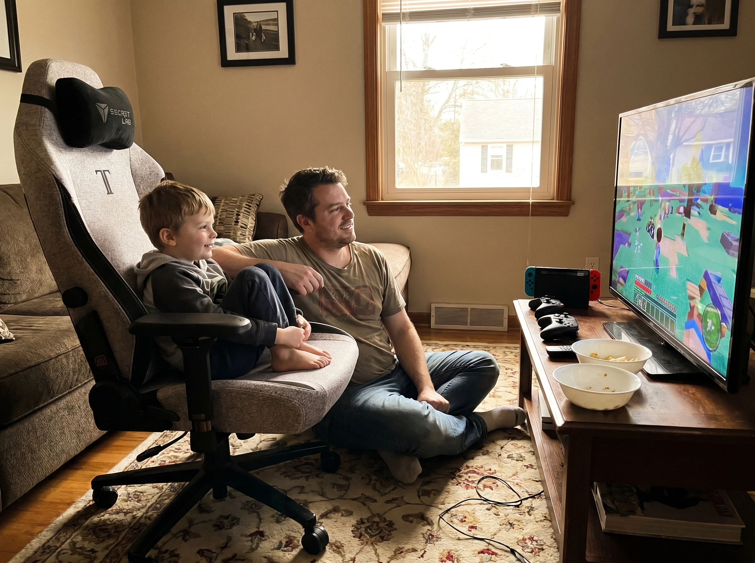 A dad sitting cross-legged on the floor next to a gaming chair, watching the screen alongside his son, both relaxed and engag