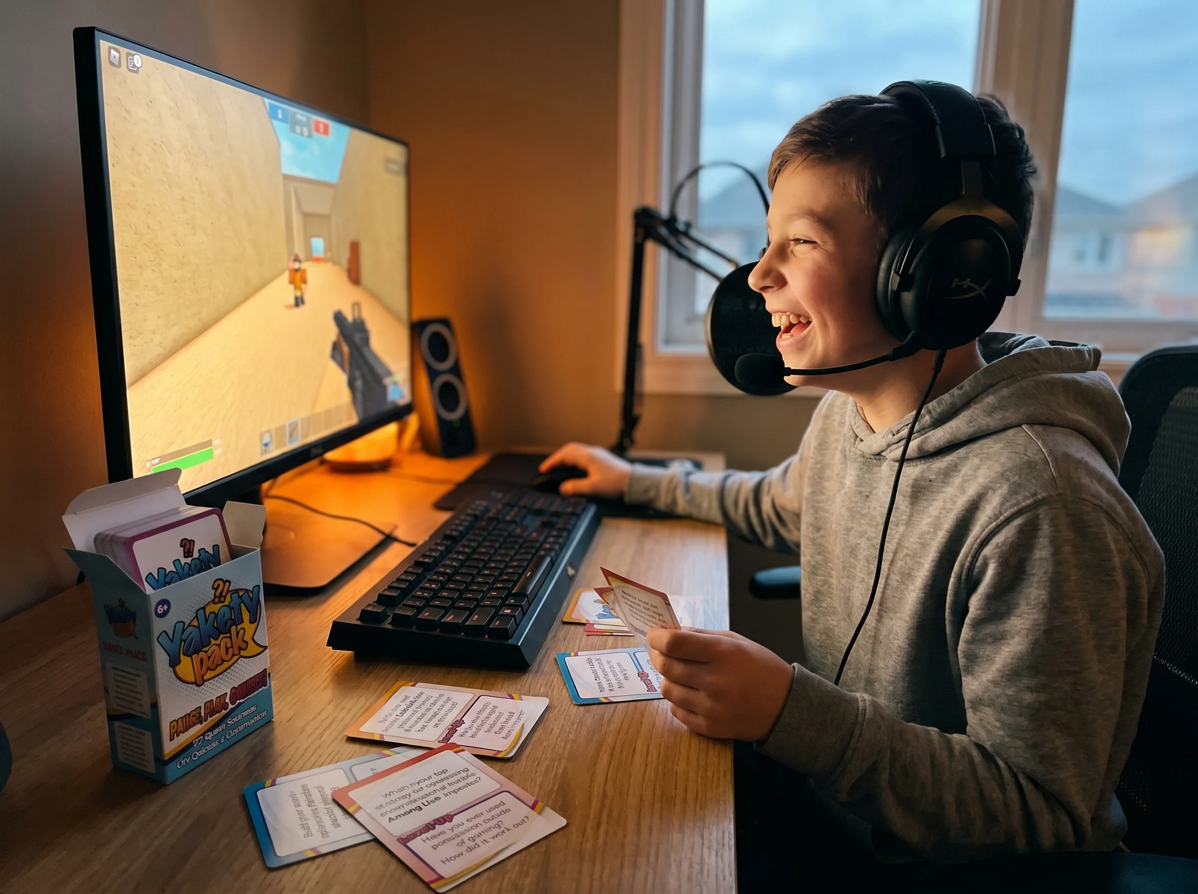 A kid in a hoodie sitting at a desk wearing headphones, laughing and talking into a mic while playing on a computer, glow fro