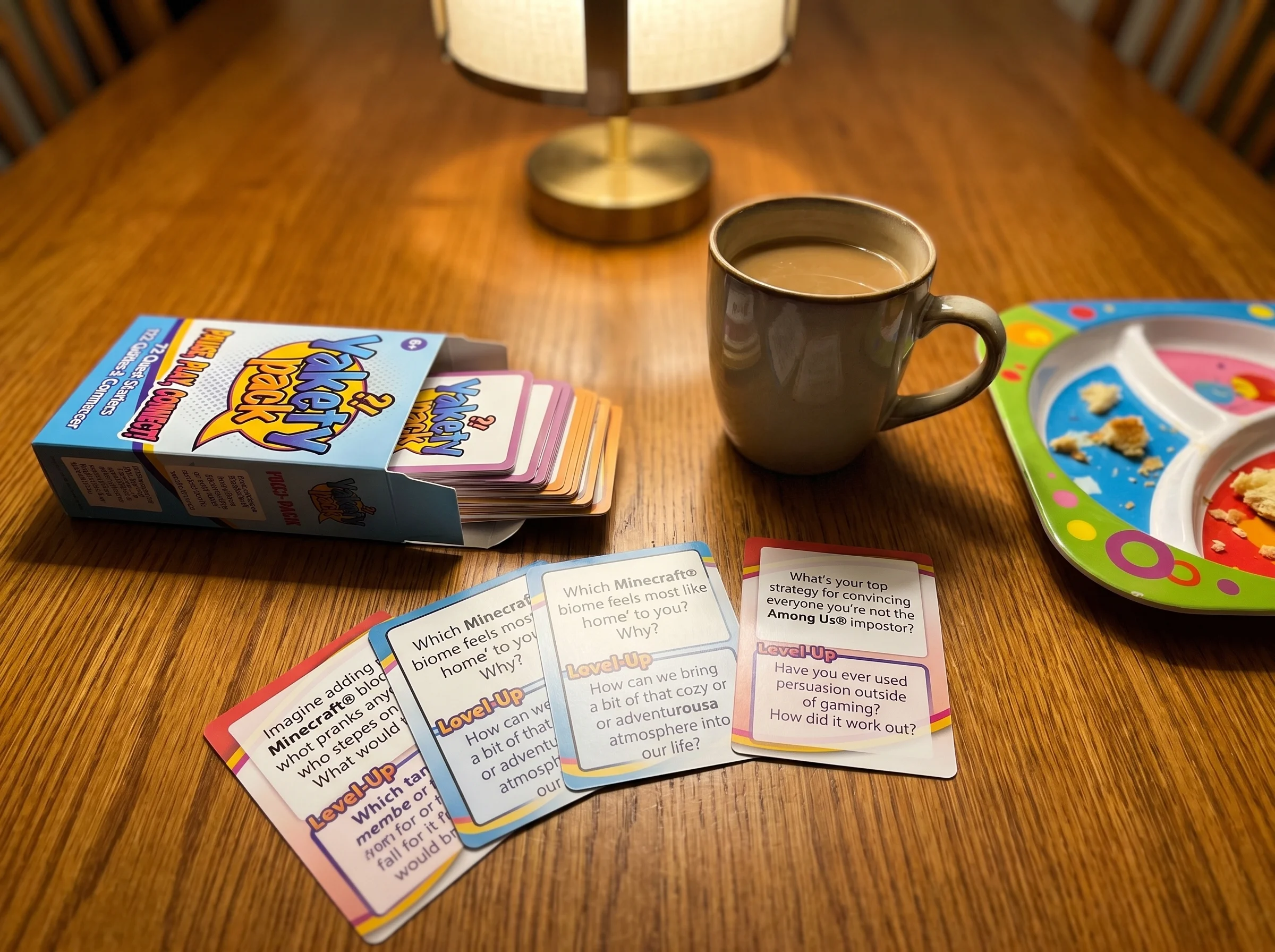 Close-up of a deck of conversation starter cards fanned out on a dinner table next to a coffee mug and a kid's plate, warm ov