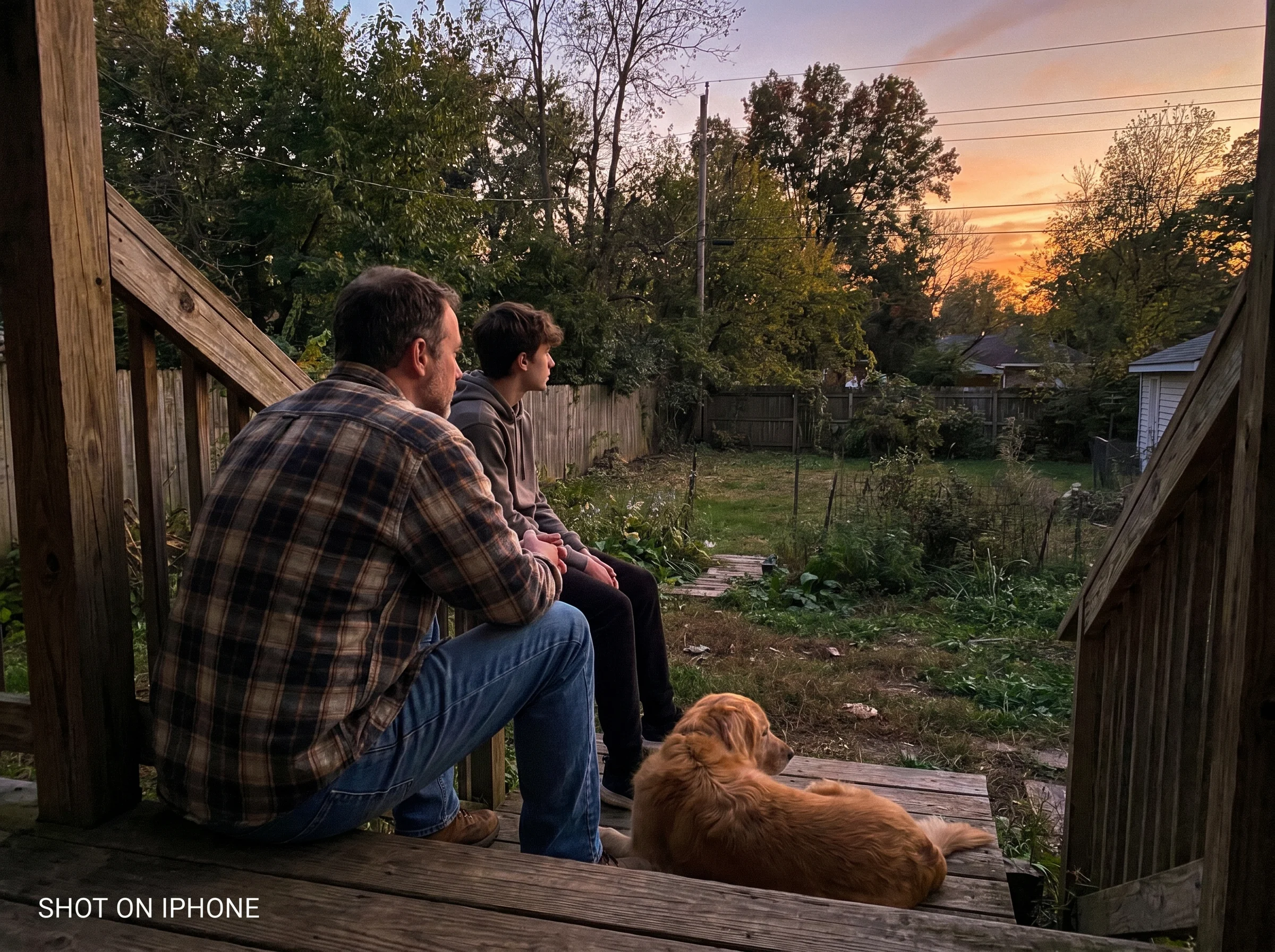 Father and teenage son sitting on a back porch step at dusk, both looking out at the yard rather than at each other, casual a