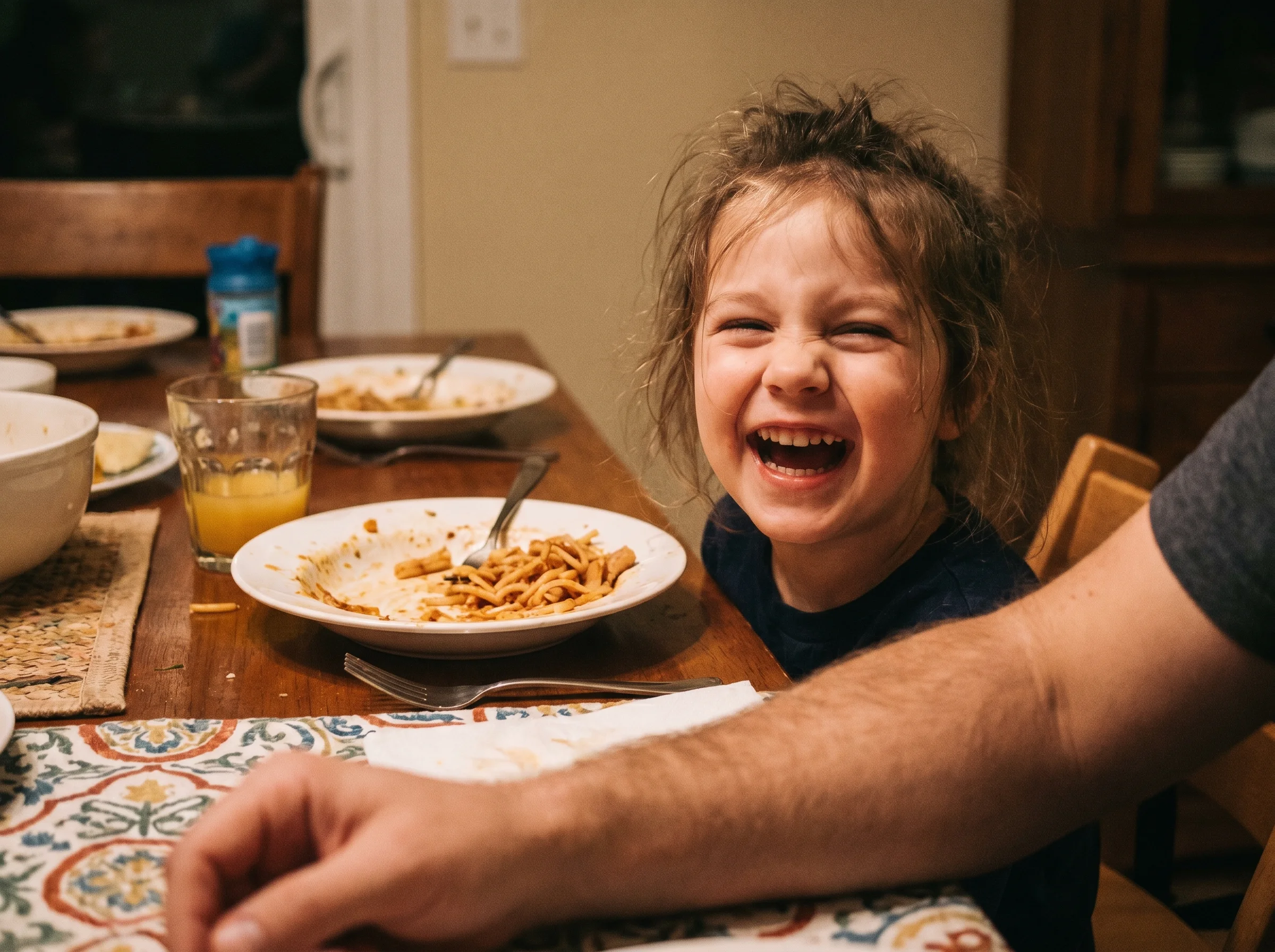 A close-up of a kid's face mid-laugh at a dinner table, eyes crinkled, real unposed joy, soft evening light