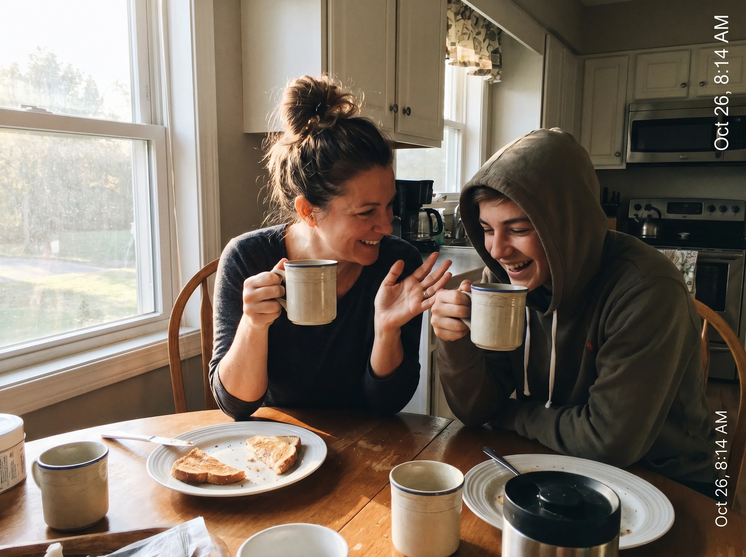 A parent and teenager at a breakfast table the next morning, mugs in hand, mid-conversation, soft morning light through a win