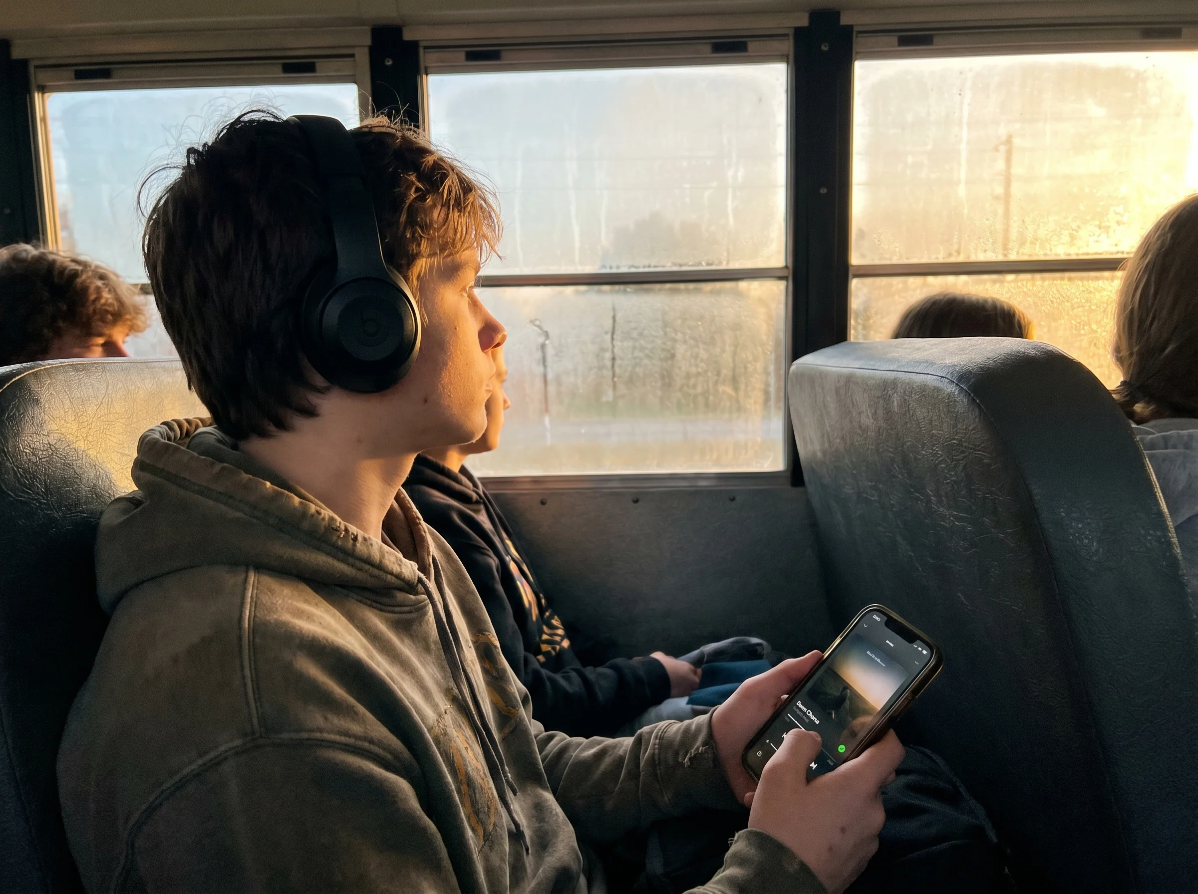 A teen wearing headphones looking out a school bus window at dawn, phone in hand showing a music app, real morning light