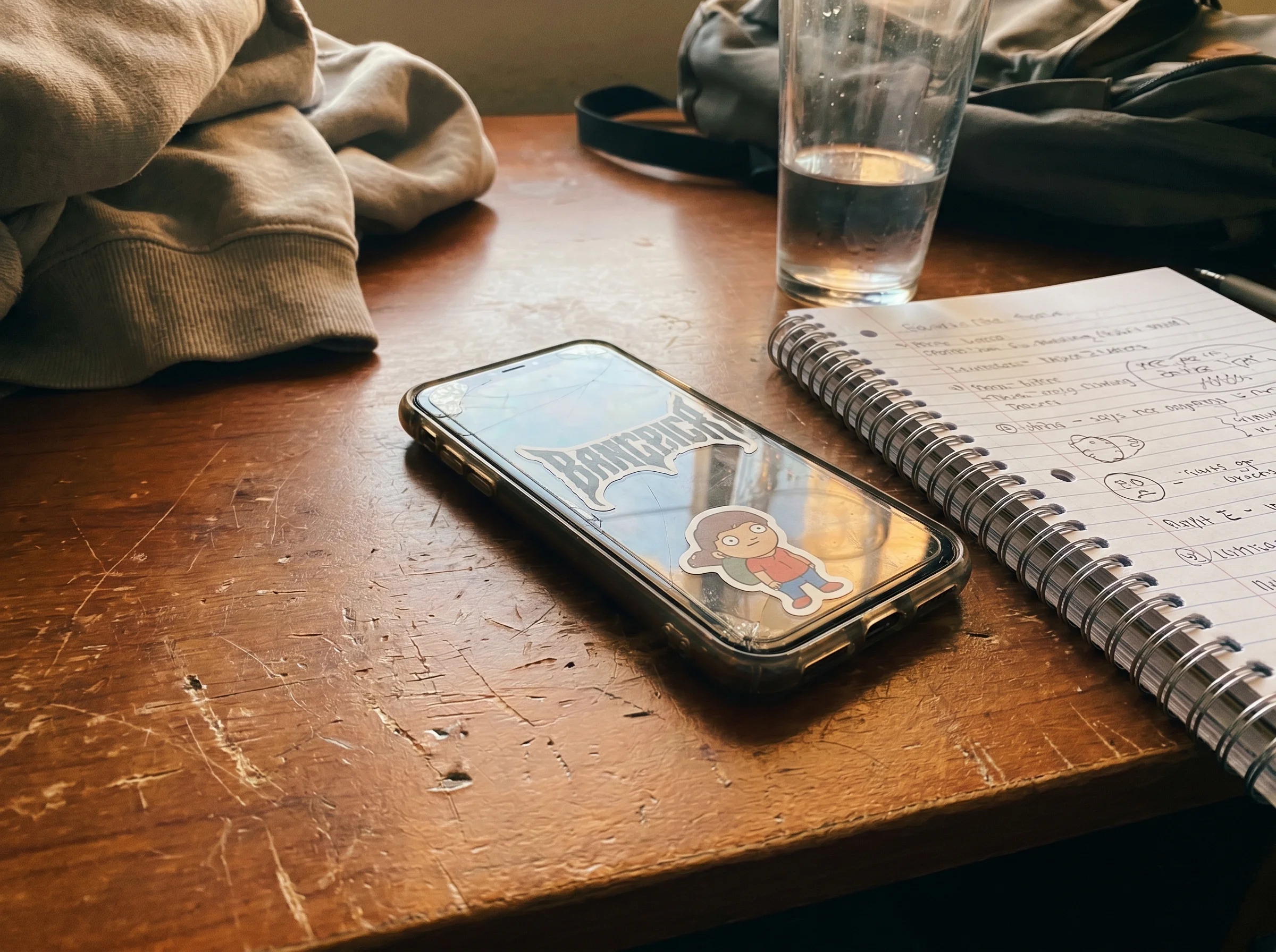 A 13-year-old's phone resting on a wooden desk next to a notebook and a half-finished glass of water, soft warm afternoon lig