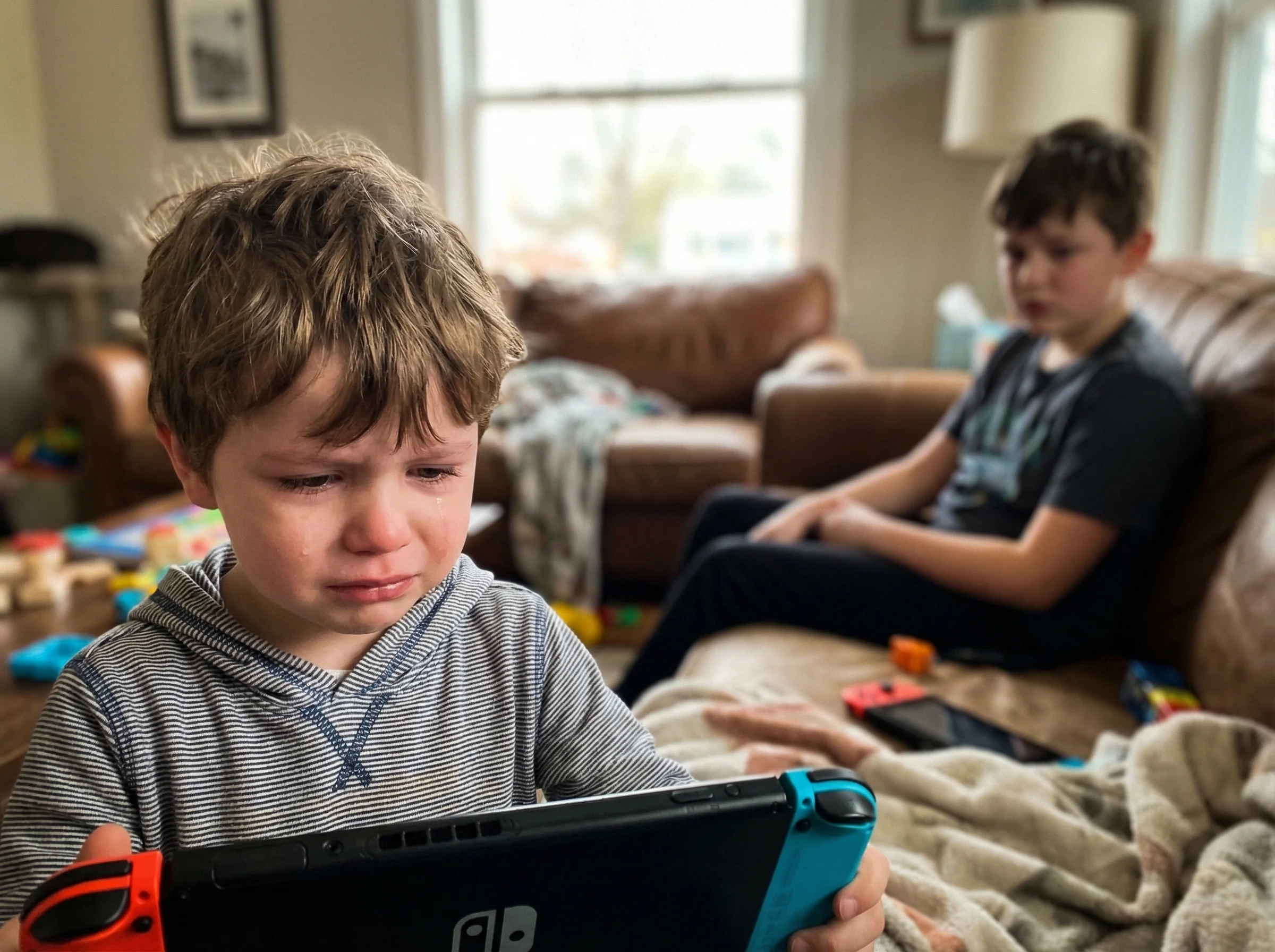 A young boy's face showing genuine distress while looking at a Nintendo Switch screen, his older brother visible in the backg