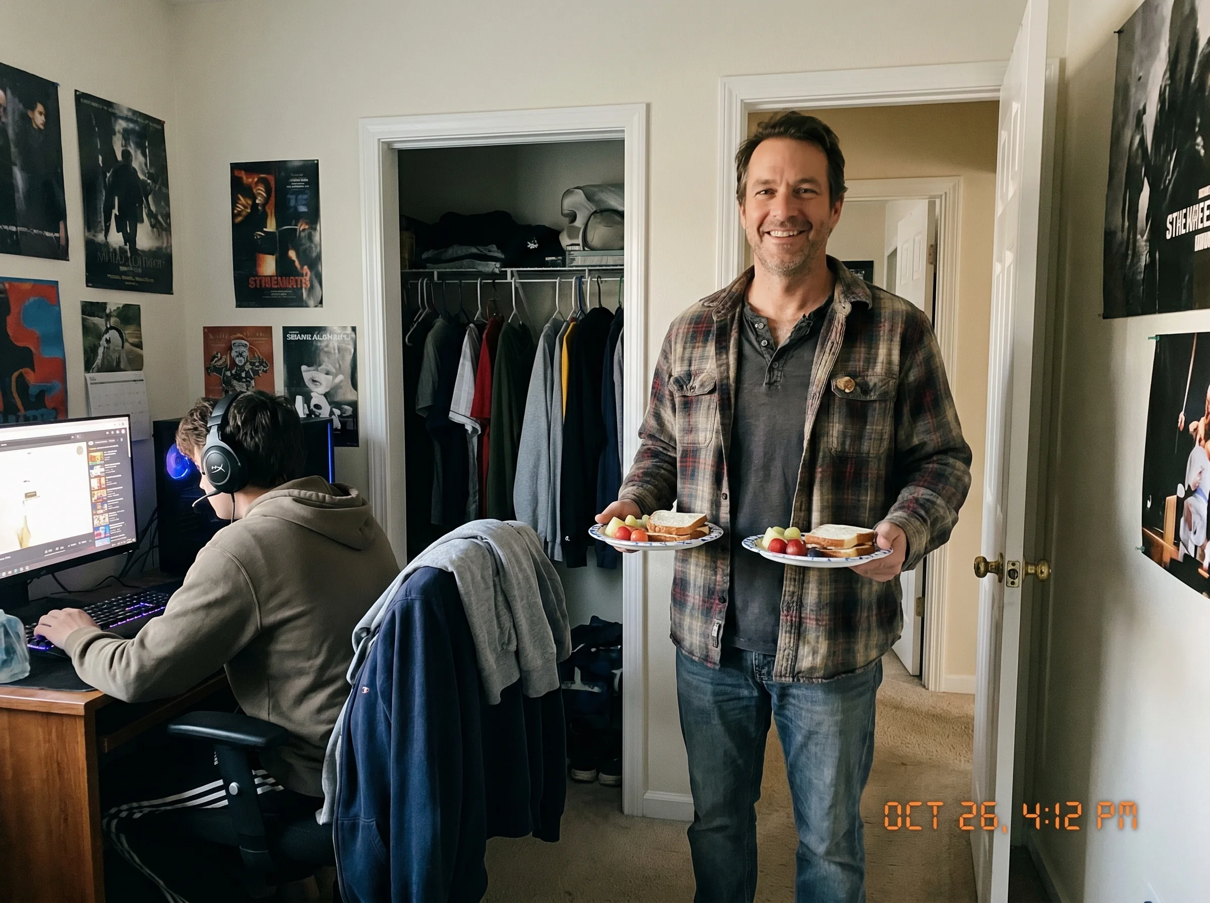 A dad standing in a doorway of a teenager's bedroom, smiling, holding two plates of snacks, kid at desk with headset on