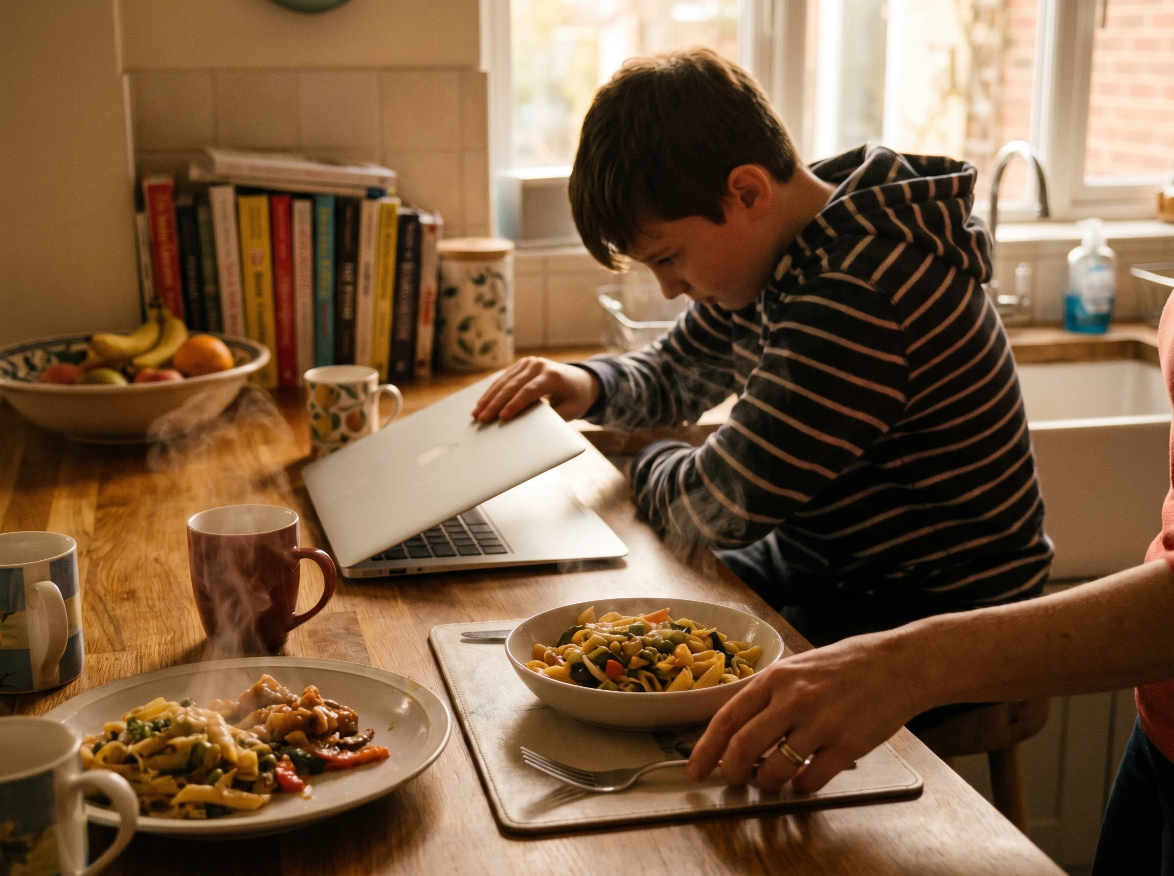 A boy closing his laptop on a kitchen counter as steam rises from dinner plates being set down, mom's hand in frame placing s